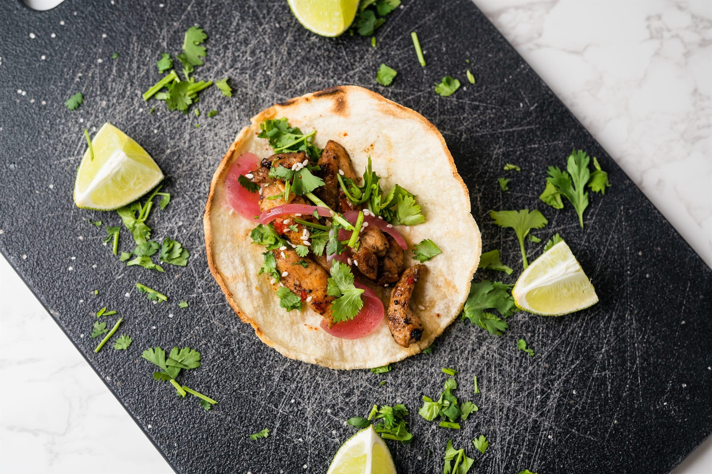 Overhead view of a street taco topped with grilled chicken, cilantro, pickled onion, and sesame seeds
